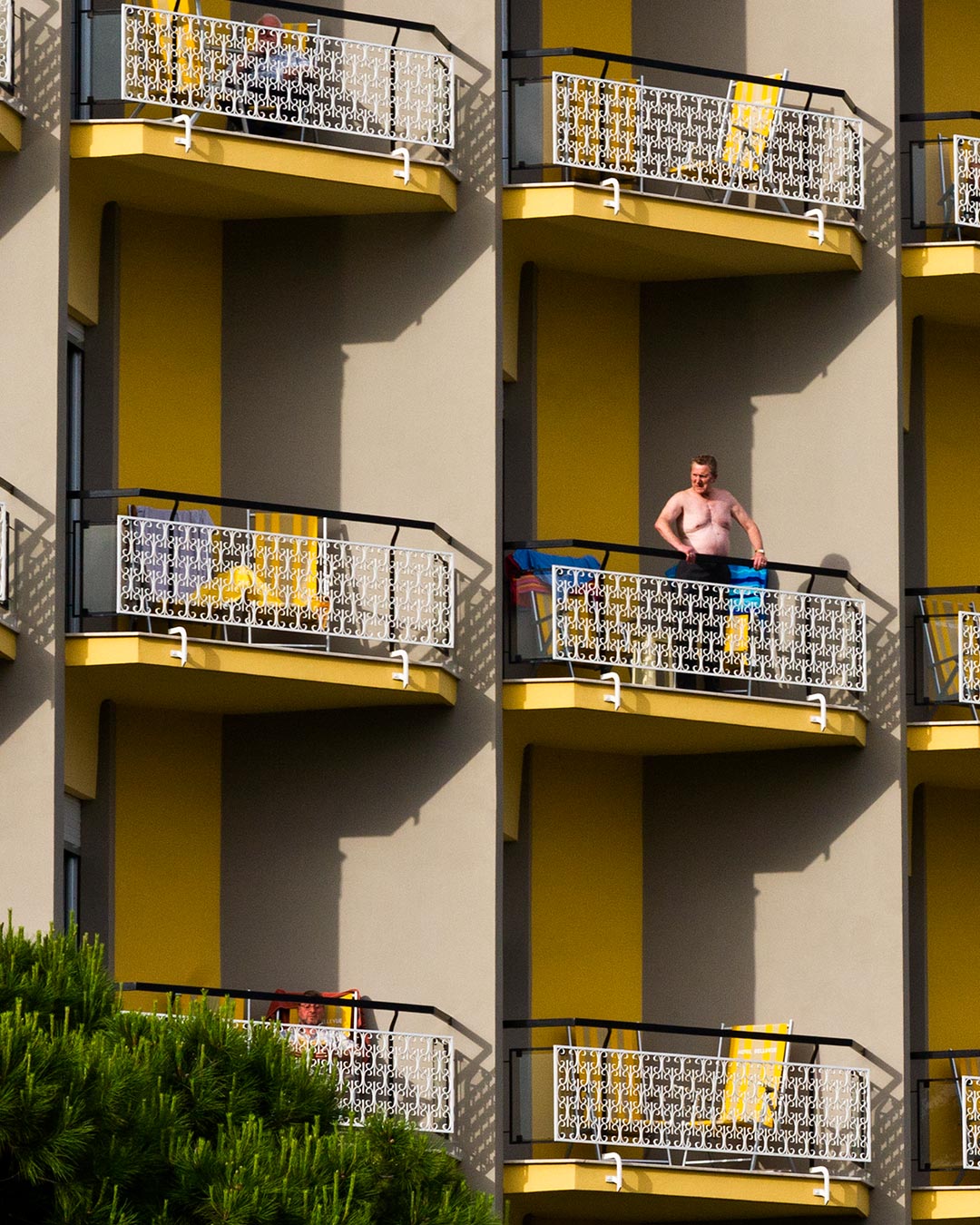 Uomo anziano a torso nudo affacciato su un terrazzo di un palazzo con molti terrazzi uguali.