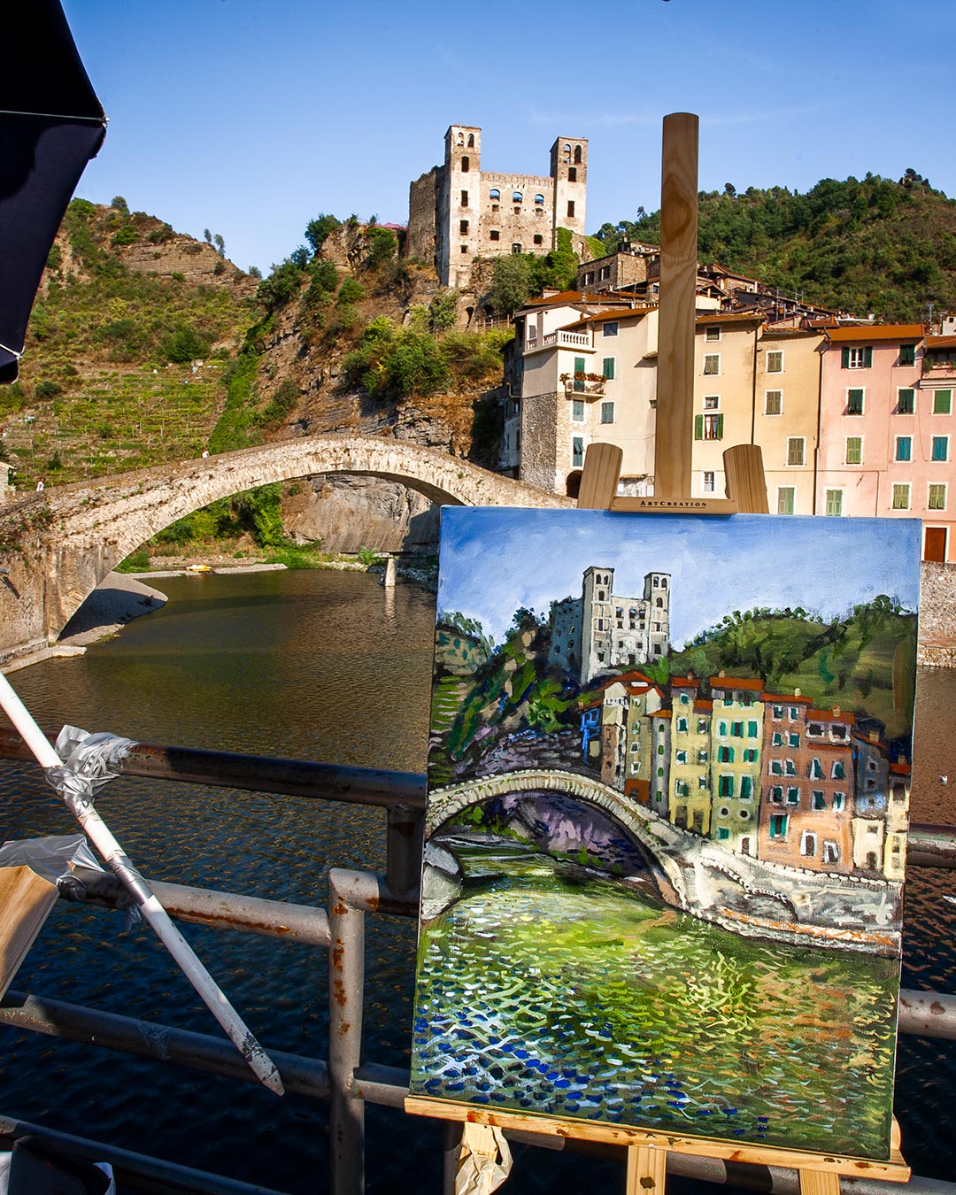 Dipinto sul cavalletto di un pittore, raffiguarante il castello e il ponte di Dolceacqua, che si trovano davanti.